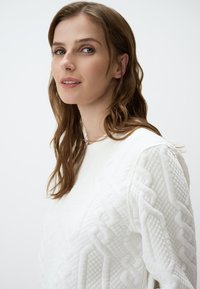 Wit gewatteerd sweater met een gestructureerd patroon en een klassieke halslijn, tentoongesteld tegen een effen witte achtergrond.