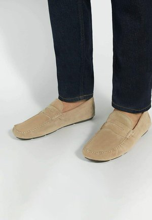 Beige suede loafers with stitched detailing and a flat black sole, paired with dark denim jeans.