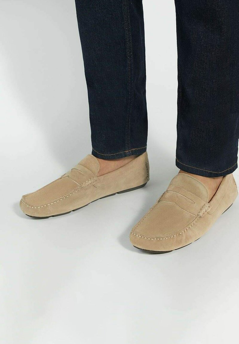 Beige suede loafers with stitched detailing and a flat black sole, paired with dark denim jeans.