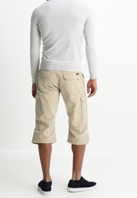 Shorts cargo beige clair avec poches latérales et ourlets, associés à une chemise ajustée à manches longues gris clair et des baskets foncées.