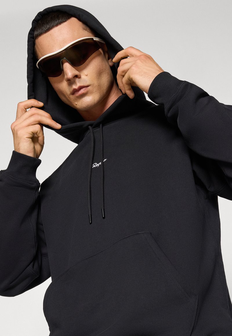 Sort bomulds-hoodie med kængurulomme, snore og hvidt broderet logo. Modellen bærer mørkt tonede solbriller.