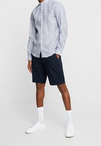 Camicetta azzurra chiara con colletto rialzato, abbinata a pantaloni corti blu navy, scarpe da ginnastica bianche e calzini bianchi, su sfondo neutro.
