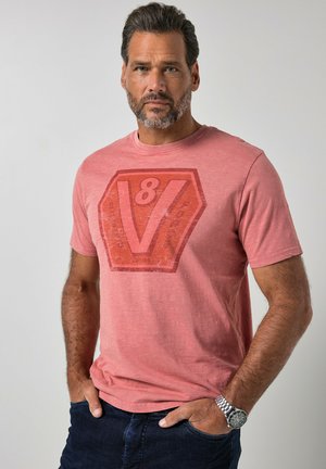 Homme aux cheveux courts et foncés avec une barbe portant un t-shirt rouge délavé avec un grand logo V8 et un jean bleu foncé, mains dans les poches, fond neutre.