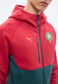 Mand iført rød og mørkegrøn lynlåstrøje med guld Puma-logo og en grøn cirkulær emblem på brystet.