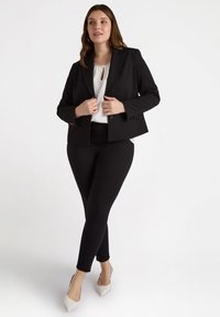 Svart blazer med en strukturerad design, parat med en vit blus med en nyckelhålsdetalj, svarta figurnära byxor och ljusa högklackade skor.