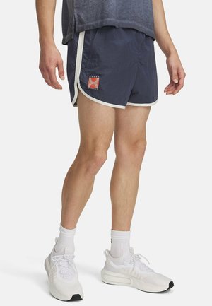 Mężczyzna ubrany w granatowe sportowe szorty z białym wykończeniem i logo Under Armour, białe skarpetki oraz białe buty do biegania, stojący na białym tle.