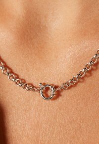 Zilveren ketting met ronde sluiting, met in elkaar grijpende schakels en een gladde textuur, die op een huidskleurige ondergrond ligt.