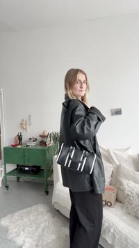 Μαύρη δερμάτινη τσάντα με λευκές λεπτομέρειες, ορθογώνιο σχήμα και επάνω χερούλια, συνδυασμένη με μια χαλαρή μαύρη ζακέτα, τοποθετημένη σε έναν φωτεινό εσωτερικό χώρο.