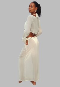 Femme aux cheveux bouclés portant un ensemble transparent blanc en deux pièces avec des ornements en forme de coquillage, se tenant pieds nus contre un fond gris uni.