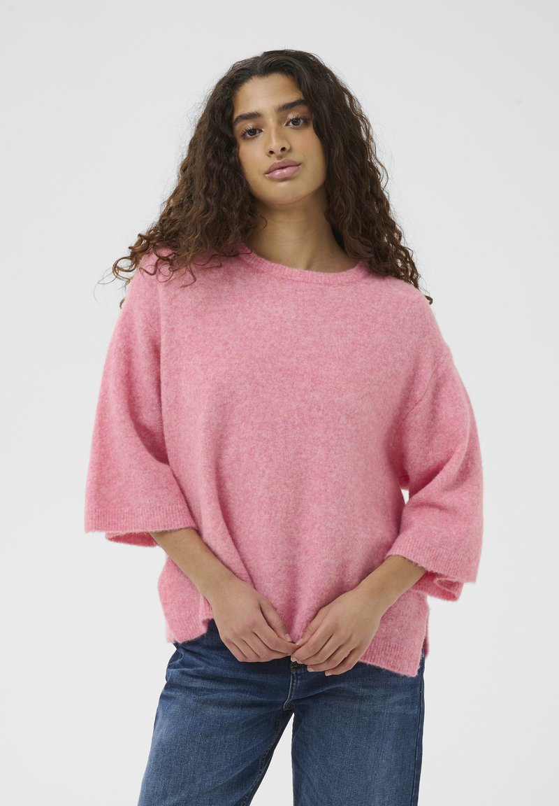 Joven mujer con cabello largo y rizado, vestida con un suéter rosa holgado y jeans azules, posando frente a un fondo claro y liso.