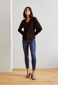 Svart långärmad blus med V-ringning, puffiga axlar, tillsammans med åtsittande blå jeans och svarta remsandaler på ett trägolv.