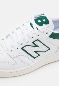 Vit sportskor i läder med gr öna accenter, en framträdande grön "N"-logotyp och en texturerad gummisula. Snören är vita.