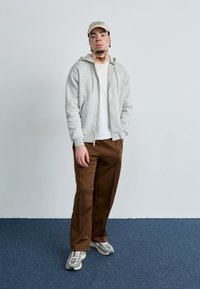 Grå hættejakke med lynlås, hvid t-shirt, brune brede bukser. Lyse beige baseballcap, grå sneakers med mesh detaljer, står på blå tæppe.