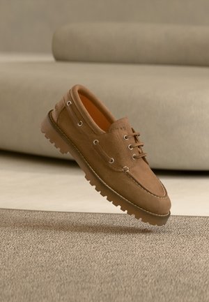 Chaussure à lacets en daim marron avec semelle épaisse rainurée sur tapis beige texturé et canapé beige flou en arrière-plan.