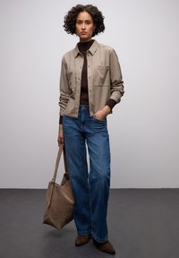 Beiges Hemd mit Knopfleiste und zwei Brusttaschen, brauner Rollkragenpullover, blaue Jeans mit weitem Bein, braune Wildleder-Stiefeletten und eine beige Tragetasche.