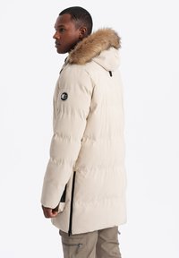 Krémová puffer bunda s kapucí podloženou kožešinou, bočními zipy a nášivkou s logem na rukávu. Prošívaná textura a délka ke kolenům.