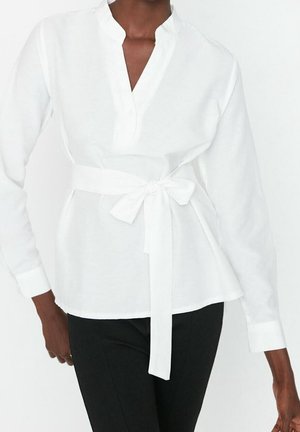 Frau trägt eine weiße Langarmbluse mit V-Ausschnitt und gebundenem Taillengürtel, kombiniert mit schwarzen Hosen vor einfachem Hintergrund.