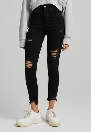 Czarne obcisłe jeansy z wysokim stanem, z przetarciami na udach i kolanach, zestawione z białymi sneakersami na platformie i szarą bluzką z długim rękawem.