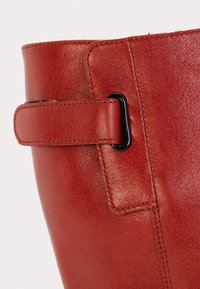 Gros plan sur la tige d'une botte en cuir rouge avec une lanière cousue attachée à travers un anneau métallique noir.