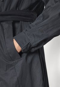 Dark grey, lightweight coat with a belted waist, featuring a visible pocket and a textured, crinkled fabric. Subtle logo detail on the sleeve.
