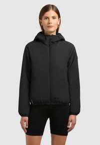 Ženska, oblečena v črno jakno z zadrgo in kapuco ter črne kolesarske hlače, stoji ob enobarvnem svetlo sivi ozadju.