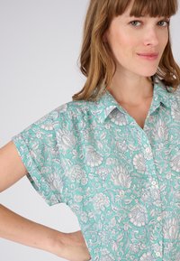 Blouse à manches courtes en turquoise clair avec un motif floral blanc. Présente un col pointu et une fermeture à boutons à l'avant.