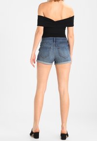 Top negro sin hombros con detalle retorcido, combinado con shorts de mezclilla azules con dobladillos enrollados y bolsillos traseros, usado con tacones negros.