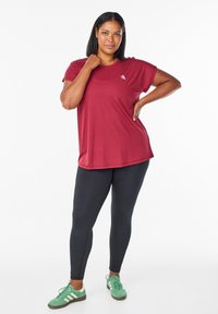 Žena stojí s jednou rukou na bedroch a druhou sa dotýka ramena, oblečená v červenom tričku, čiernych legínach a zelených teniskách.