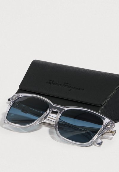 γυαλιά ηλίου με διάφανο πλαίσιο και σκούρους μπλε φακούς, τοποθετημένα πάνω σε μια μαύρη υφασμάτινη θήκη με ανάγλυφο λογότυπο.