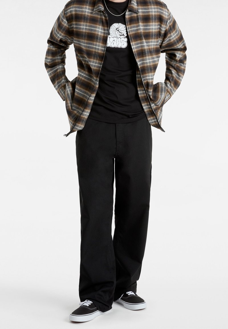 Personne portant une veste zippée à carreaux marron sur un t-shirt graphique noir, un pantalon large noir et des baskets noires avec des semelles blanches.