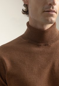 Hombre viste un suéter de cuello alto marrón acanalado, mostrado desde los hombros hasta debajo de la nariz, contra un fondo beige liso.