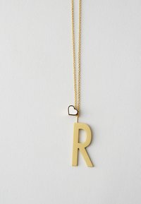Collier doré mettant en avant un grand pendentif en forme de lettre "R" avec un petit accent en forme de cœur, fabriqué en métal avec une texture lisse.