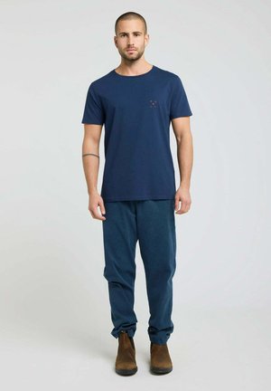 Homme debout portant un t-shirt bleu marine, un pantalon ample bleu foncé et des bottes en daim marron sur un fond gris clair uni.