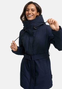 Parka isolante bleu marine avec un col haut, une ceinture, une poche zippée et des cordons de serrage réglables. Tissu texturé avec une finition lisse.