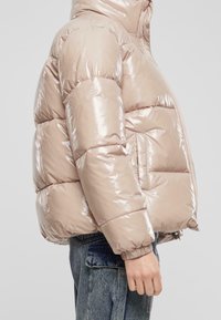 Kurtka puchowa w błyszczącym, jasnobeżowym materiale z kapturem, wyposażona w zamek błyskawiczny i boczną kieszeń, pikowany wzór oraz elastyczne mankiety.