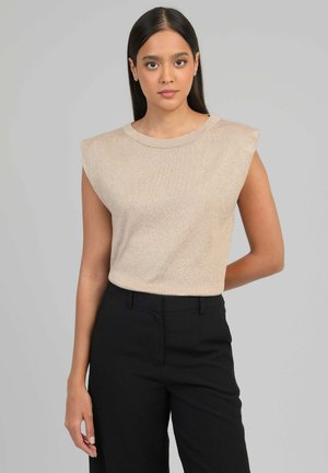 Femme aux longs cheveux foncés portant un haut beige sans manches avec épaules rembourrées et un pantalon noir, debout devant un fond gris uni.