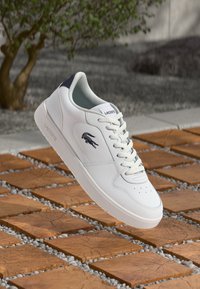 Sapatilha de couro branca com uma textura suave, detalhe azul-marinho no calcanhar e bico perfurado. Apresenta o logótipo da Lacoste na lateral.