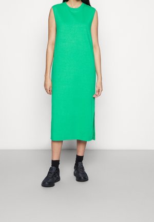 Mouwloze groene midi-jurk met zijsplitten, gemaakt van een zachte gebreide stof. Gecombineerd met zwarte sneakers en sokken. Eenvoudige, rechte silhouet.