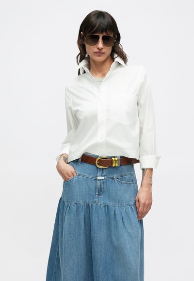 Biela košeľa s gombíkmi a vyhrnutými rukávmi, doplnená vrstvenou rifľovou sukňou. Hnedý opasok so zlatou prackou a veľké slnečné okuliare.