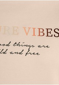 Camiseta de un suave color beige claro que presenta un texto multicolor que dice "PURE VIBES" y un guion que dice "las cosas buenas son salvajes y libres". Textura suave.