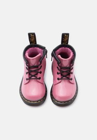 Γυαλιστερά ροζ παπουτσάκια για νήπια με μαύρα κορδόνια, μαύρες σόλες και κίτρινα ράμματα, φωτισμένα από πάνω σε λευκό φόντο.