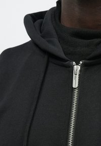 Camisola preta com capuz e fecho de correr à frente, com tecido texturizado e gola alta. O fecho de correr prateado adiciona detalhe ao design.