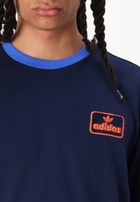 Marinblå skjorta med en blå krage, prydd med ett texturerat orange Adidas-logotypmärke på bröstet. Mjuk tyg och avslappnad passform.