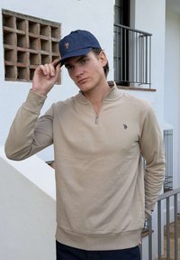 Jeune homme ajustant une casquette bleu marine, portant un pull beige à fermeture éclair quart, avec un petit logo, debout près d'un bâtiment blanc avec une fenêtre et une rambarde en métal.