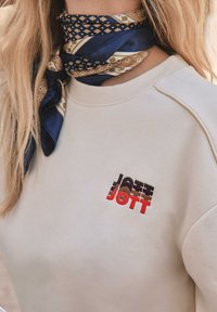 Sudadera de color beige claro con un logo bordado en relieve en rojo y marrón. Llevada con una bufanda de patrón azul y marrón alrededor del cuello.