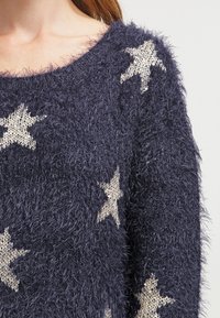 Pullover blu navy in maglina morbida con scollatura ampia e motivi a stella beige. La texture è morbida e leggermente pelosa.