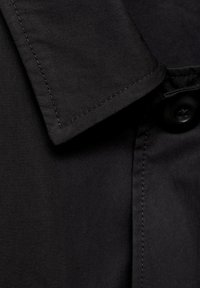 Zwarte stoffen shirt met een spitse kraag, enkele knoopsluiting en een gladde textuur met zichtbare stikseldetails.