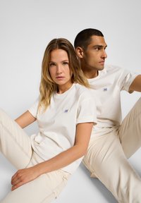 T-shirt in cotone beige con maniche arrotolate, caratterizzato da un piccolo logo blu, abbinato a pantaloni in cotone beige chiaro con cuciture a contrasto.