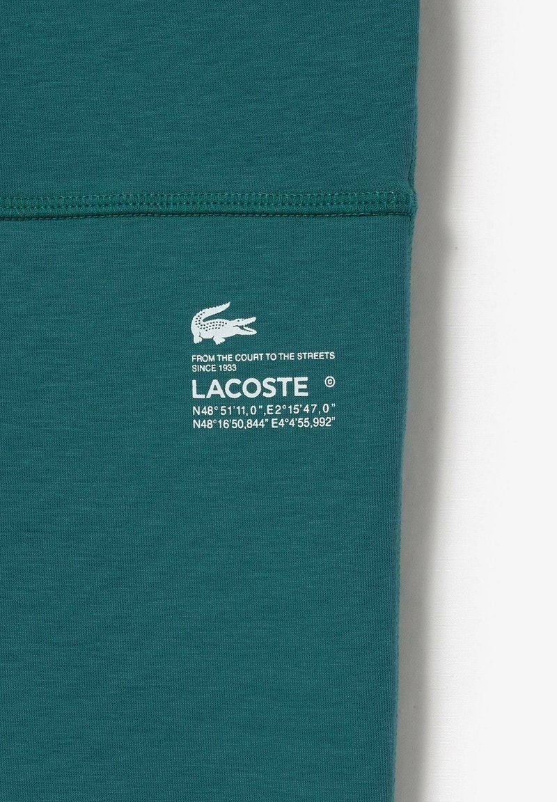 Turkusowa koszulka bawełniana z białym logo Lacoste i tekstem. Gładka tekstura, przeszyte szwy oraz dopasowany krój.