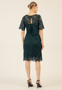 Tüllkleid in Teal mit kurzen Ärmeln, floralen Mustern, Rückenschlüssellochdetail, mittellanger Schnitt und Schlitz am Saum. Kombiniert mit schwarzen High Heels.